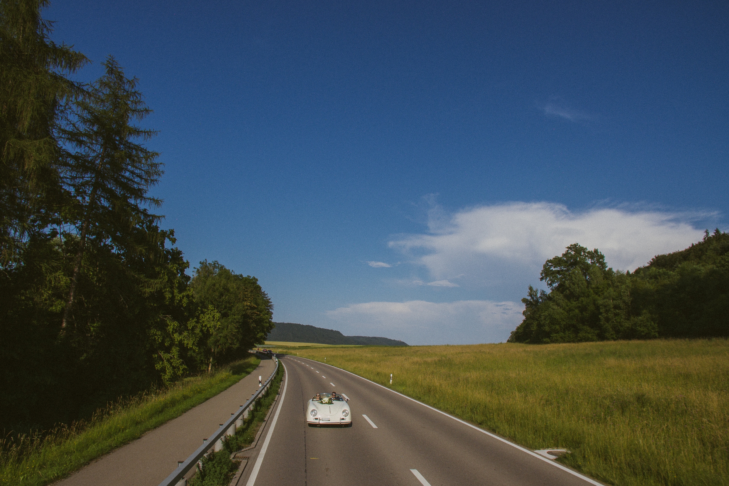 Hochzeit in der Schweiz: Brautpaar fährt mit dem Oldtimer über eine Landstraße, kein weiteres Auto weit und breit