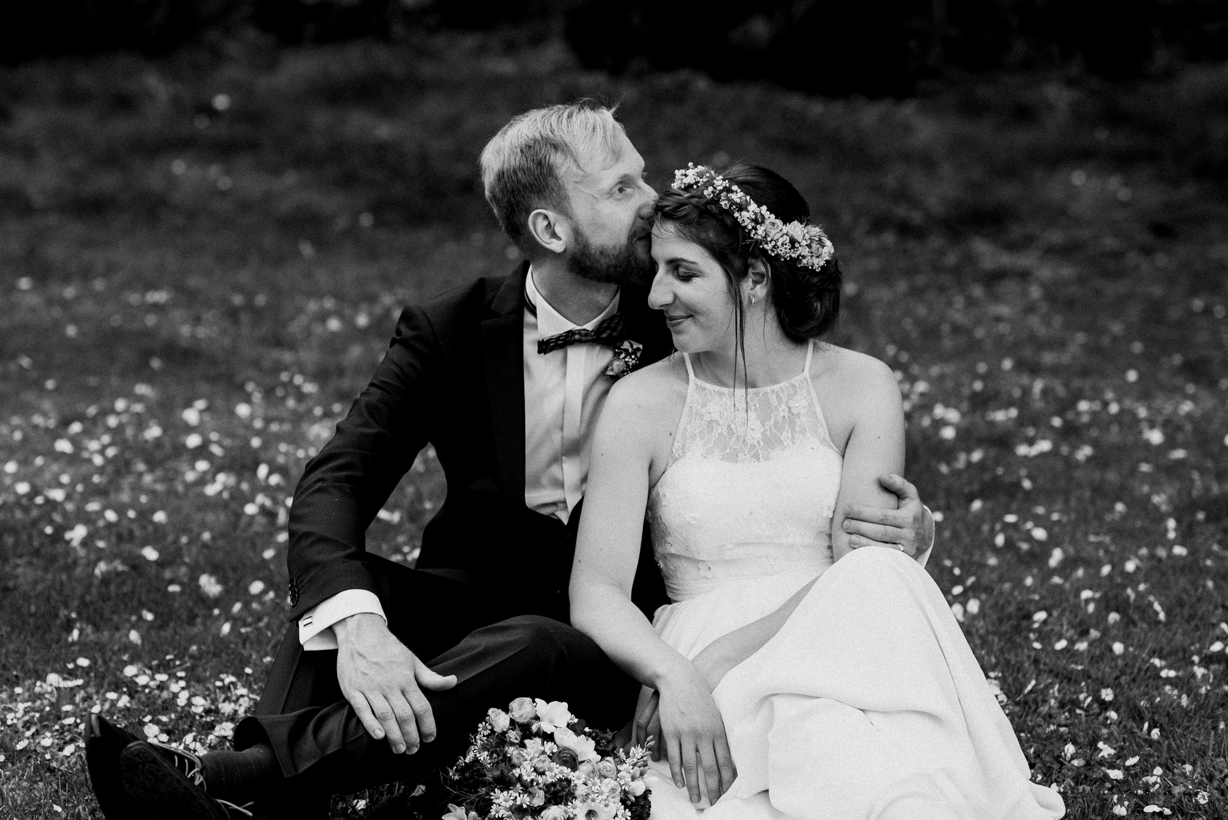Outdoor-Hochzeit: Brautpaar sitzt im Gras, Bräutigam küsst Braut auf die Stirn
