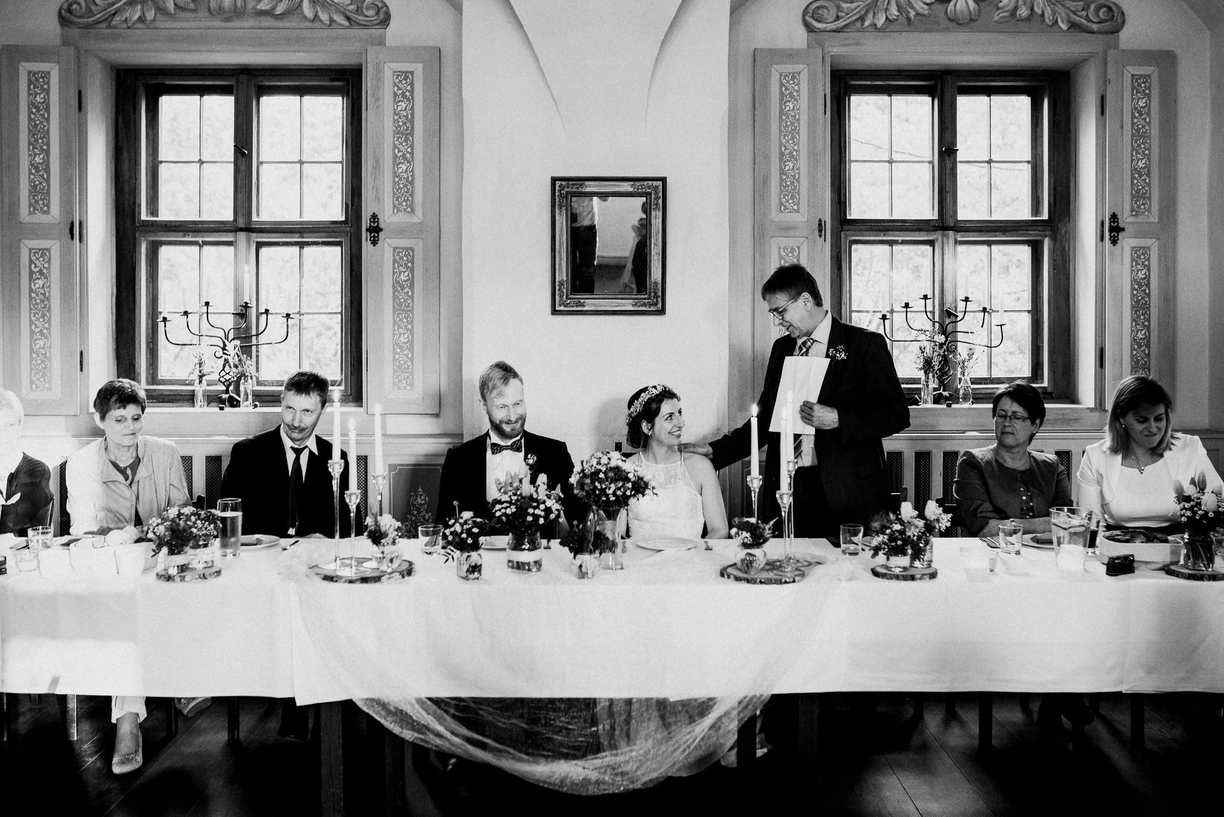 Outdoor-Hochzeit: Brautpaar an einer Tafel, Hochzeitsgast hält eine Rede und beugt sich zur Braut