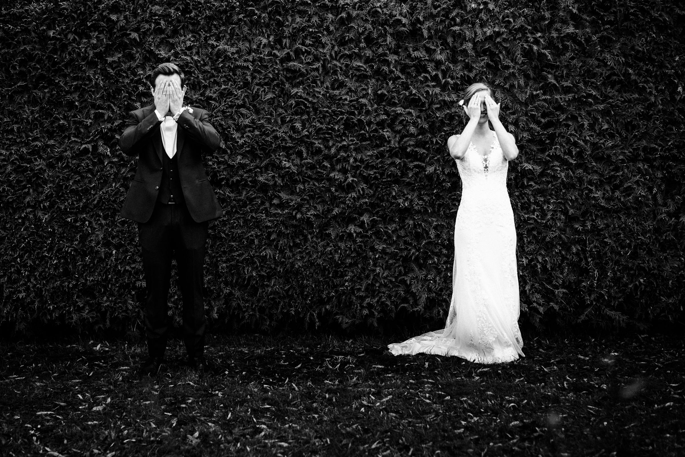 das Brautpaar der Silvester-Hochzeit steht nebeneinander vor einer efeubewachsenen Wand und hält sich die Augen zu