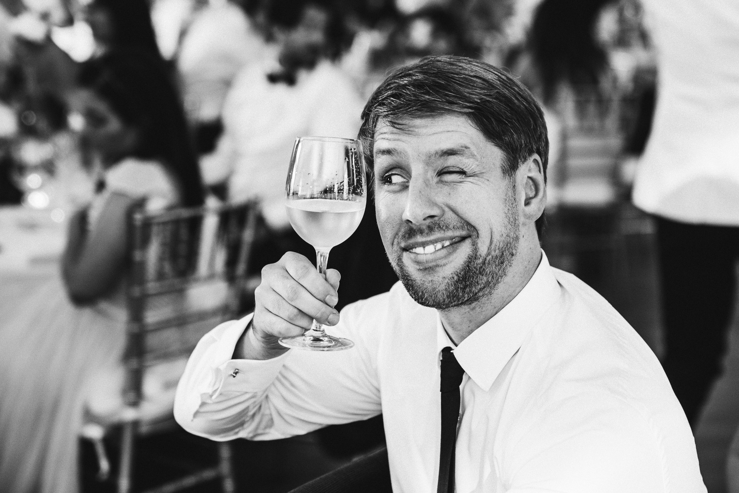 Septemberhochzeit: Hochzeitsgast zwinkert durch Weinglas hindurch dem Brautpaar zu