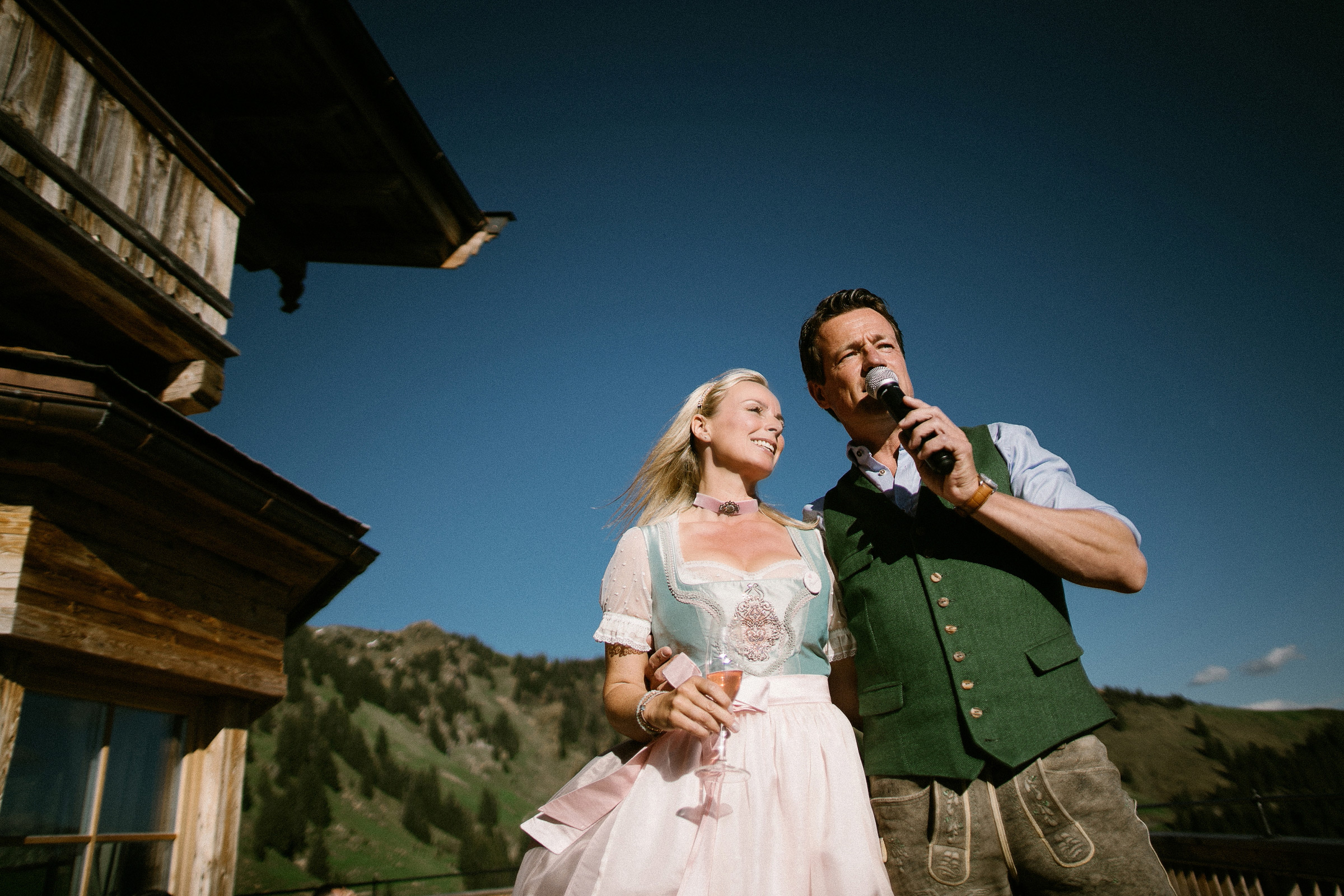 Alpenhochzeit: Weitwinkelaufnahme des Brautpaars, das auf der Terrasse eine Rede hält