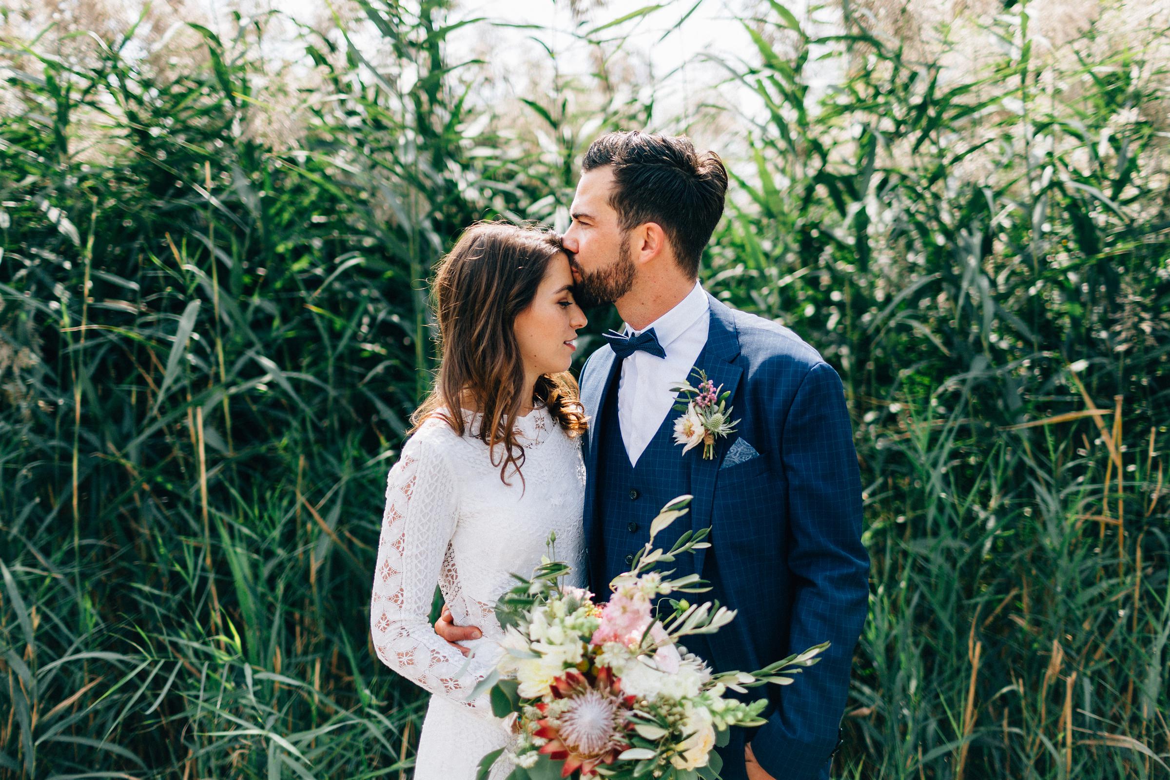 das Braupaar der mediterranen Hochzeit eng umschlungen vor einer Wand aus hohem Gras