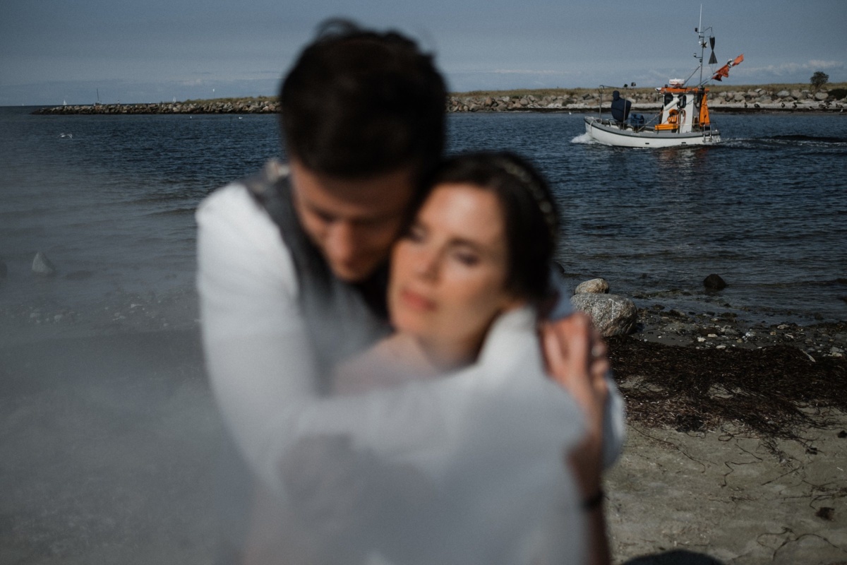 Strandhochzeit in Kiel: Portraitaufnahme des Brautpaares, mit Fokus auf den vorbeifahrenden Kutter.