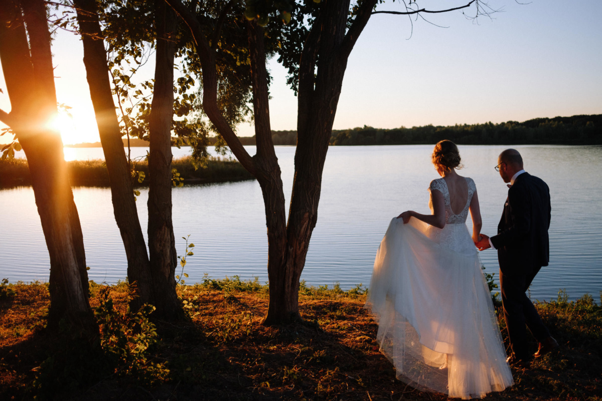 Vintage-Hochzeit: Brautpaar läuft im Abendlicht Hand in Hand an den See