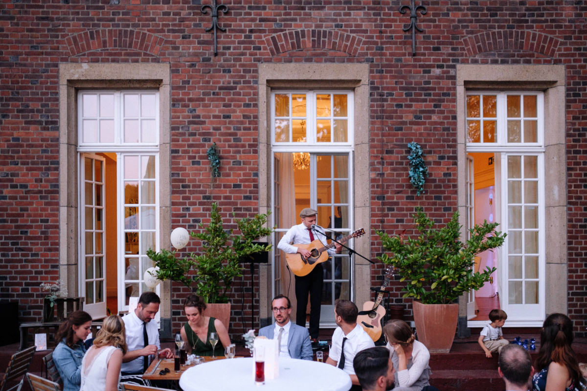 Vintage-Hochzeit: Gäste sitzen draußen, im Hintergrund ein Gitarrenspieler
